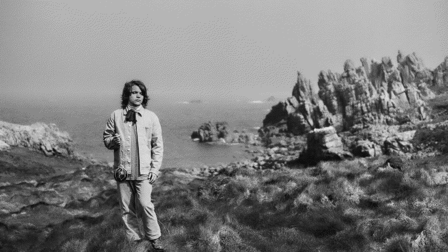 Black and white image of a young man standing on the grassy foreground with the sea and cliffs behind him. He holds a piece of rope from which dangles an orb.