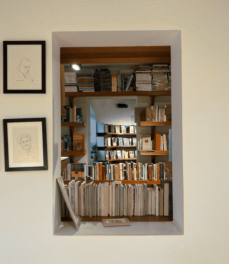 A view of a blank wall within a bookshop that has an interior window through which books and shelves filled with books in the next room are visible, to the left are two framed hand-drawn portraits.
