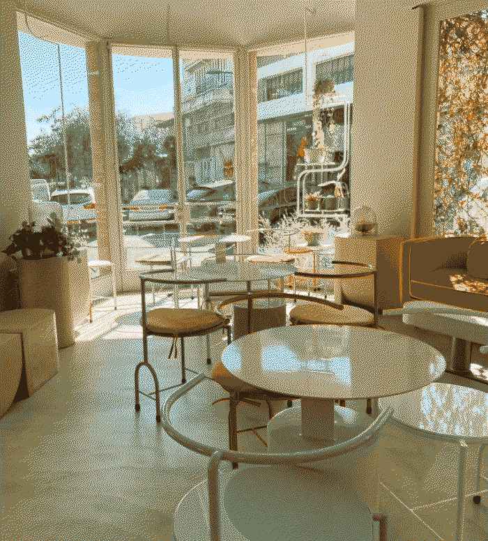 View of an empty café with tables and chairs in the foreground and the street through large windows in the background. A palette of browns and blues.