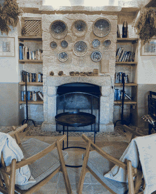 Stone fireplace surrounded by books and ceramic plates and two armchairs facing it in the foreground.