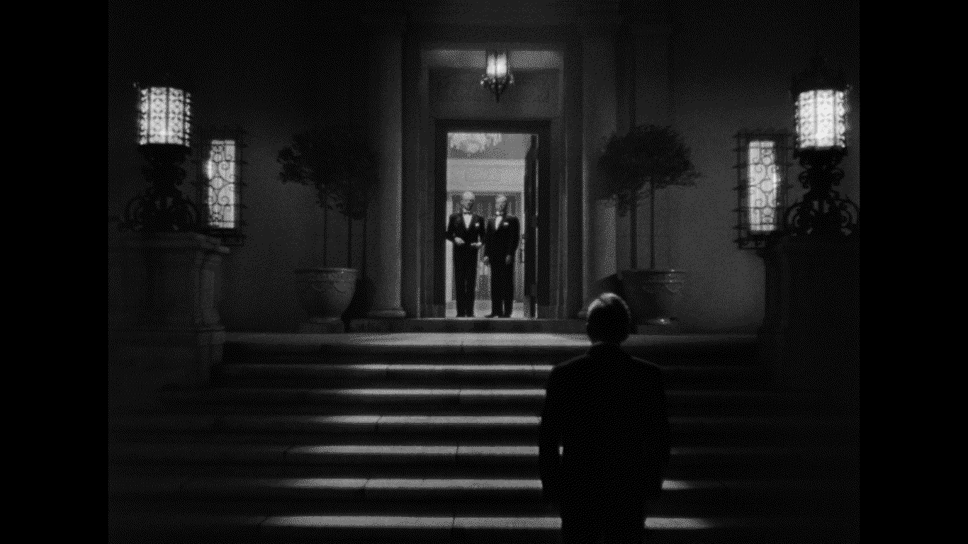 Two men in black tie suits stand at an open doorway of large mansion and look down at a third man facing away from the camera.
