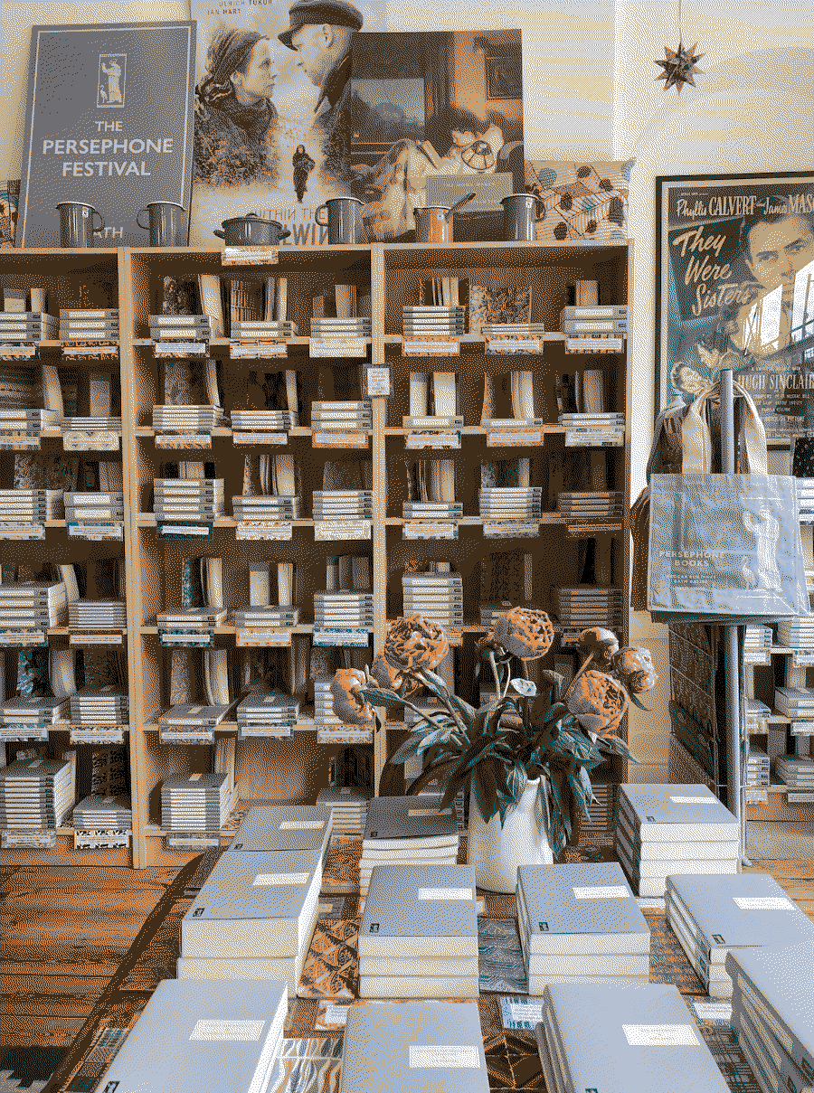 Indoor scene of a bookshop with books in shelves in the background and more books on the table in the foreground and a vase with flowers.